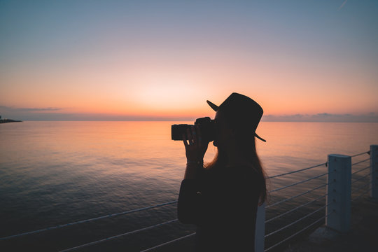 Silhouette of woman photographer in hat with digital camera during taking photos of sea at pier at sunset - Powered by Adobe