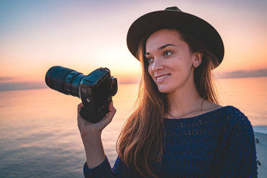 Portrait of happy young woman traveler photographer in hat with dslr camera during taking photos of sea at sunset