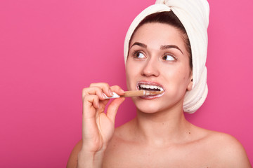 Close up portrait of young adorable woman standing isolated over pink studio background and brushing her teeth, wearing white towel, having thoughtfull facial expression, dreams about something.