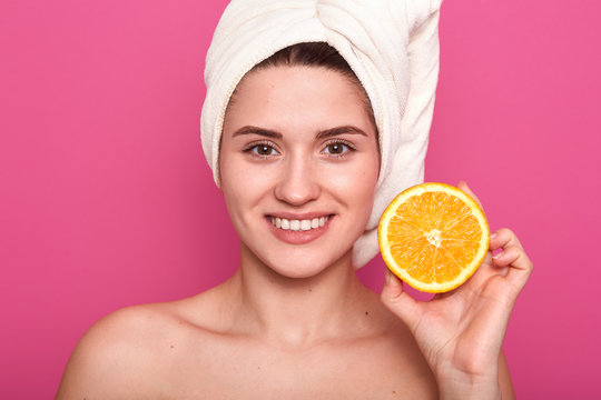 Picture of adorable woman with half of orange in her hand, wrapped white towel on her head, looking smiling directly at camera, posing isolated over pink studio background. Skin care concept.