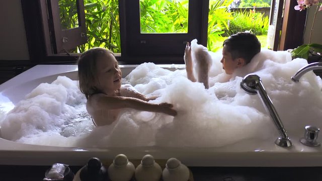 Kids taking bubble bath. Children playing with soap foam in a large white bath and big window with tropical green garden in Ubud, Bali, Indonesia.