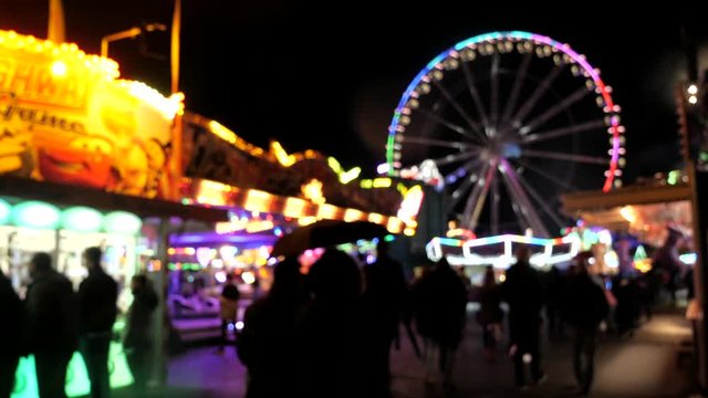 Bokeh blur shot showing people walking over funfair during rainy day at night