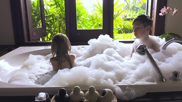 Child girl and boy taking bath with shampoo and soap. Kids playing with foam in bathroom with nature view outside window.
