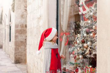 Christmas shopping. Little child girl on market street outside. Funny kid in santa hat looks at on...