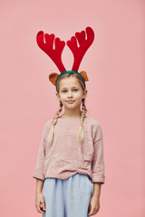 Portrait of cute little girl wearing deer antlers and looking at camera over pink background