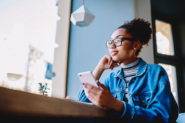 Happy hipster girl in stylish spectacles spending time with smartphone device in cafeteria connected to wireless 4g internet, positive woman dreaming about future life while holding telephone in hand