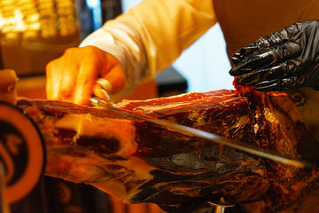 Hands of a woman in black gloves cut a slice of traditionally Spanish jamon de bellota in the restaurant or market or store.