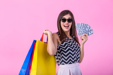 Photo of cheerful elegant young woman in sunglasses holding fan of money and colorful shopping bags, looking aside, isolated on pink background