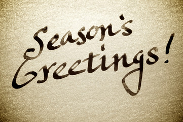 Handwritten Season's Greetings! holiday card message in fresh black calligraphy ink on textured sparkling gold paper