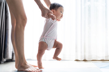 Fototapeta na wymiar Asian baby taking first steps walk forward on the soft mat. Happy little baby learning to walk with mother help at home. Mother teaching how to walk gently. Baby growth and development concept.