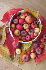 Little red and yellow apples in a white bowl on wooden table with red and yellow leaves. Autumn background