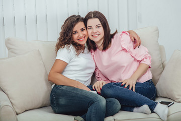 Two amazing girls sitting on the sofa, hugging one another, smiling at the camera.