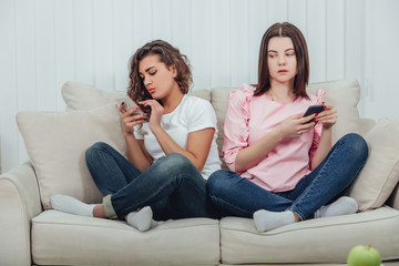 Two attractive girlfriends sitting on the sofa side by side in lotos position, with smartphones in...