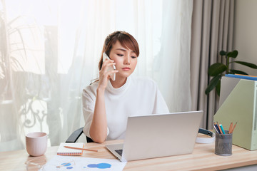 Young Asian woman using her cellphone while using laptop and working from home