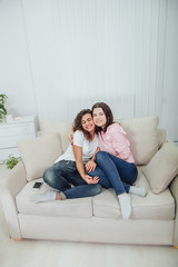 Fototapeta na wymiar Two amazing girls sitting on the sofa, hugging one another, smiling lovely, looking at the camera.