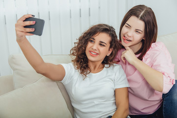 Cute young girlfriends sitting on the sofa, taking selfie, posing, making faces. Brunette girl is showing her tongue.
