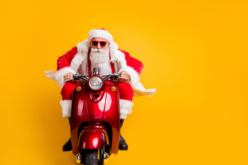 Portrait of serious hipster santa claus in red hat drive his scooter hurry rider ride to x-mas christmas party wear shirt suspenders isolated over yellow color background