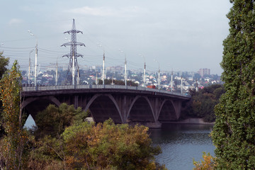 Concrete arched bridge over the reservoir in the city center. Cars move along the bridge, an electric pole with wires is visible behind the bridge.
