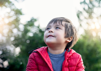 Candid shot of happy little kid looking up with smiling face sitting in the park,Cropped shot of child boy looking up to sky relaxing on his own with blurry natural green forest backgroud