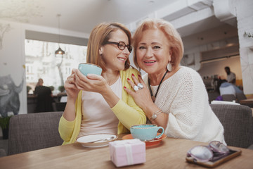 Cheerful senior woman smiling to the camera, hugging her adult daughter at the coffee shop