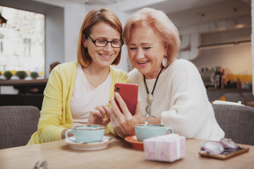 Cheerful mature woman and her lovely senior mother browsing online together, using smart phone