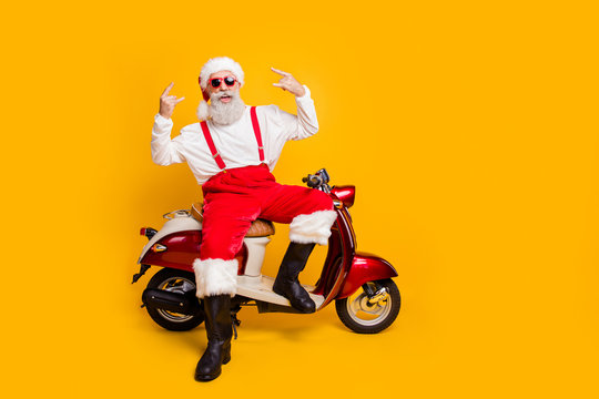 Full body photo of crazy white hair santa in festive mood riding x-mas party sitting on bike showing horns wear sun specs pants cap shirt boots isolated yellow color background