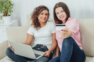 Blurred happy female friends sitting on the sofa using laptop and credit card for shopping online.