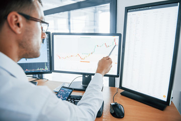 With phone and pen. Analysing graphs. Young businessman in formal clothes is in office with multiple screens. Conception of exchange and money