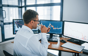 Holds cup of drink. Analysing information. Young businessman in formal clothes is in office with multiple screens. Conception of exchange and money