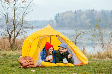 Cute young couple of millennial or generation z adventurers or travellers sit in front of tent, girl makes selfie,man looks into distance contemplating and relaxing. Scandinavian lifestyle inspiration