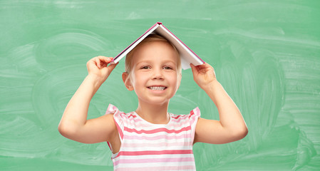 school, education and knowledge concept - smiling little girl with roof of book on top of her head...