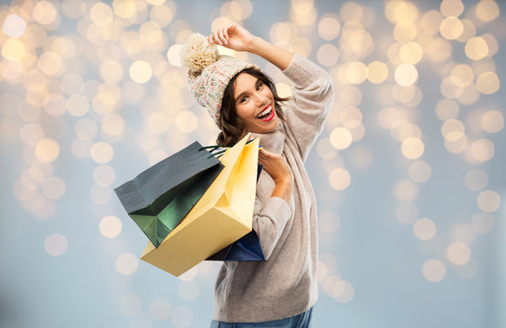 christmas, seasonal sale and consumerism concept - happy smiling young woman in knitted winter hat and sweater with shopping bags over festive lights background