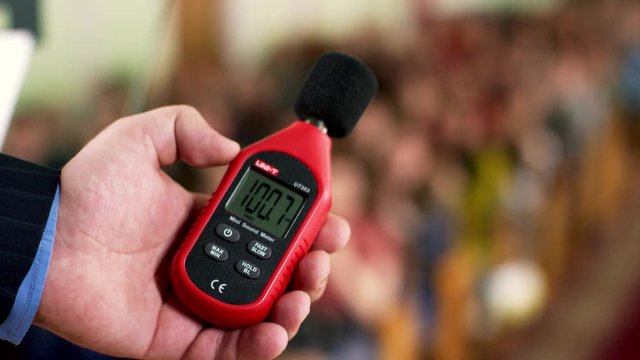 Close up of a man hand holding a red sound level meter and analyzer on clapping audience background. Stock footage. Man measuring the noise level on blurred crowd background.