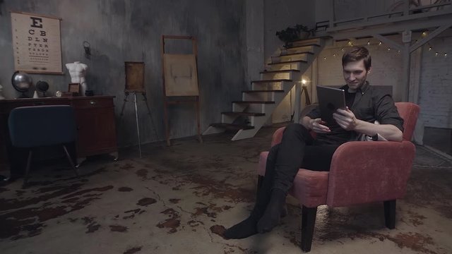 Handsome young man in red armchair using a digital tablet. Stock footage. Man looking at his device in authentic duplex art studio with grey walls.