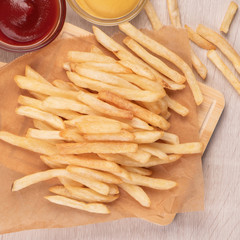 Golden yummy deep French fries on kraft baking sheet paper and serving tray to eat with ketchup and yellow mustard, top view, lifestyle.