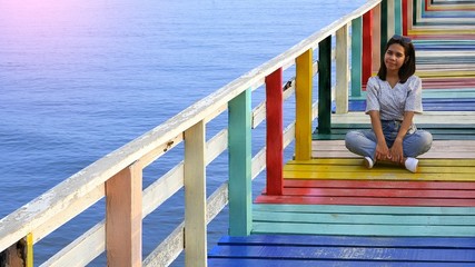 Young Asian female tourist sitting and relaxing on multicolored wooden bridge at sea viewpoint in her weekend, lifestyle in travel concept