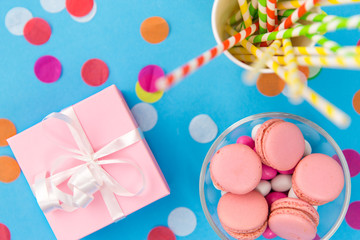 birthday party, celebration and decoration concept - gift box, pink macarons, paper straws and confetti on blue background