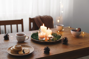 Cozy evening tea party by candlelight. Served table: cup of tea, cookies, tray, candles, pine cone, branches, led garland lights. Table setting. Christmas or New year holiday decorations. Table decor.