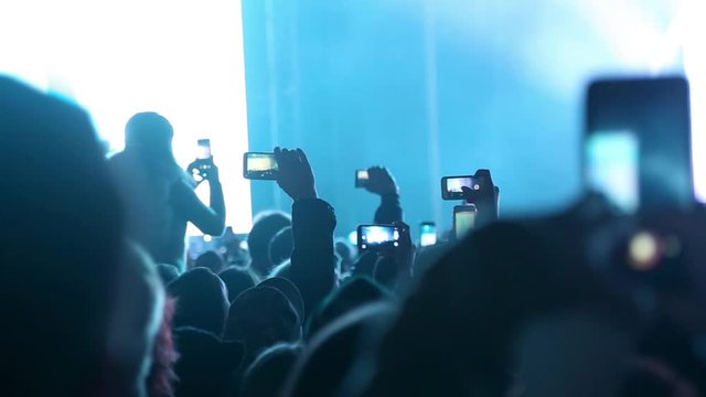 Outdoor night rock, pop concert. People cheer move lift and clap their hands in unison against the strobing stage lights. People taking video and photos on mobile smart phone at concert party crowd.