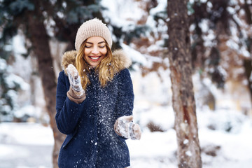 Beautiful girl playing with snow in winter forest. Smiling girl in a blue jacket and knitted hat and mittens having fun with snow falling in hands. Fashion young woman in the winter park. Christmas. 