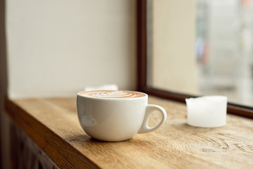 White cup of coffee with milk or cocoa with a pattern on milk foam on a wooden windowsill.