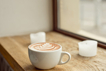 White cup of coffee with milk or cocoa with a pattern on milk foam on a wooden windowsill.