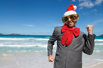 Excited businessman in Santa hat, sunglasses and big red Christmas bow dancing on a tropical beach