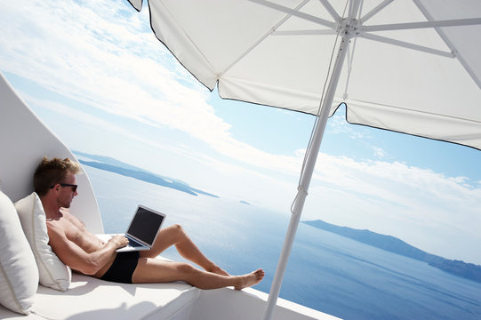 Man relaxing with laptop computer on white balcony with scenic view of the Mediterranean Sea and Santorini caldera