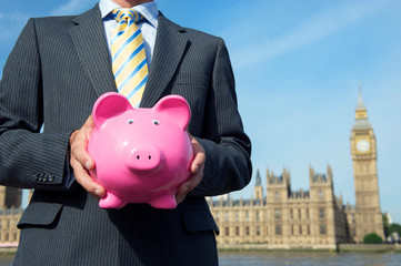 British politician holding a classic pink piggy bank in front of the Houses of Parliament in London, UK
