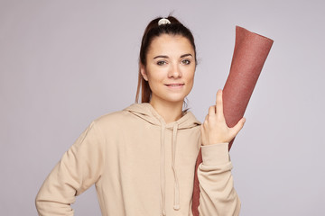 Positive attractive female trainer with perfect fresh skin biting lower lip, inviting clients at new opened gym, wearing casual comfortable hoodie, posing on grey blank studio wall for advertising.