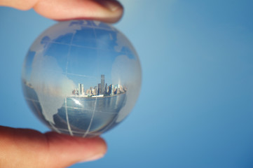 City skyline reflecting in crystal ball globe of the world held up between fingers against blue sky
