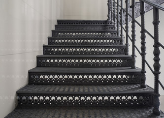 Antique metal staircase with black richly decorated wrought-iron steps and railings. Interior detail in loft style. Close-up front view, copy space. Stairs lead upward