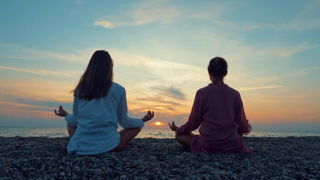 Two young women practice yoga on a sandy beach by the sea or ocean at sunset. Self-knowledge and healthy lifestyle concept.