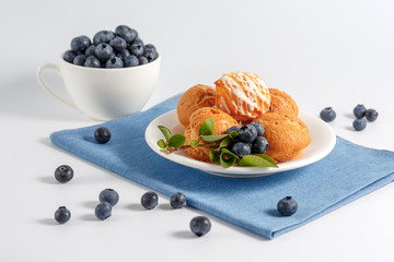 There is a delicious dessert-custard cakes eclairs with sweet filling and fresh blueberries. A white Cup filled with blueberries and cakes lying in a white plate on a blue napkin. Selective focus.